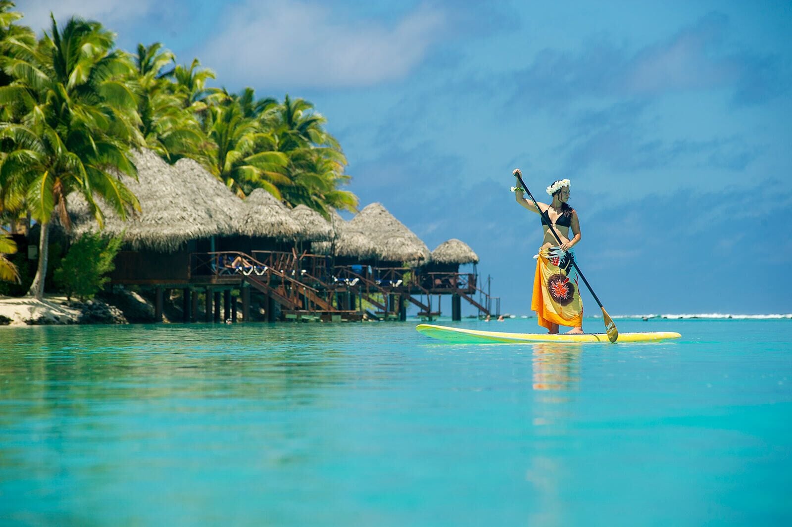 Une personne fait du paddle sur une eau turquoise, avec en arrière-plan une structure au toit de chaume et des palmiers.