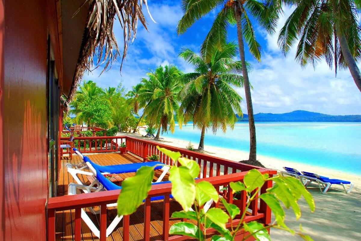 A wooden deck with blue lounge chairs and a red railing overlooking a beach with palm trees and clear blue water.