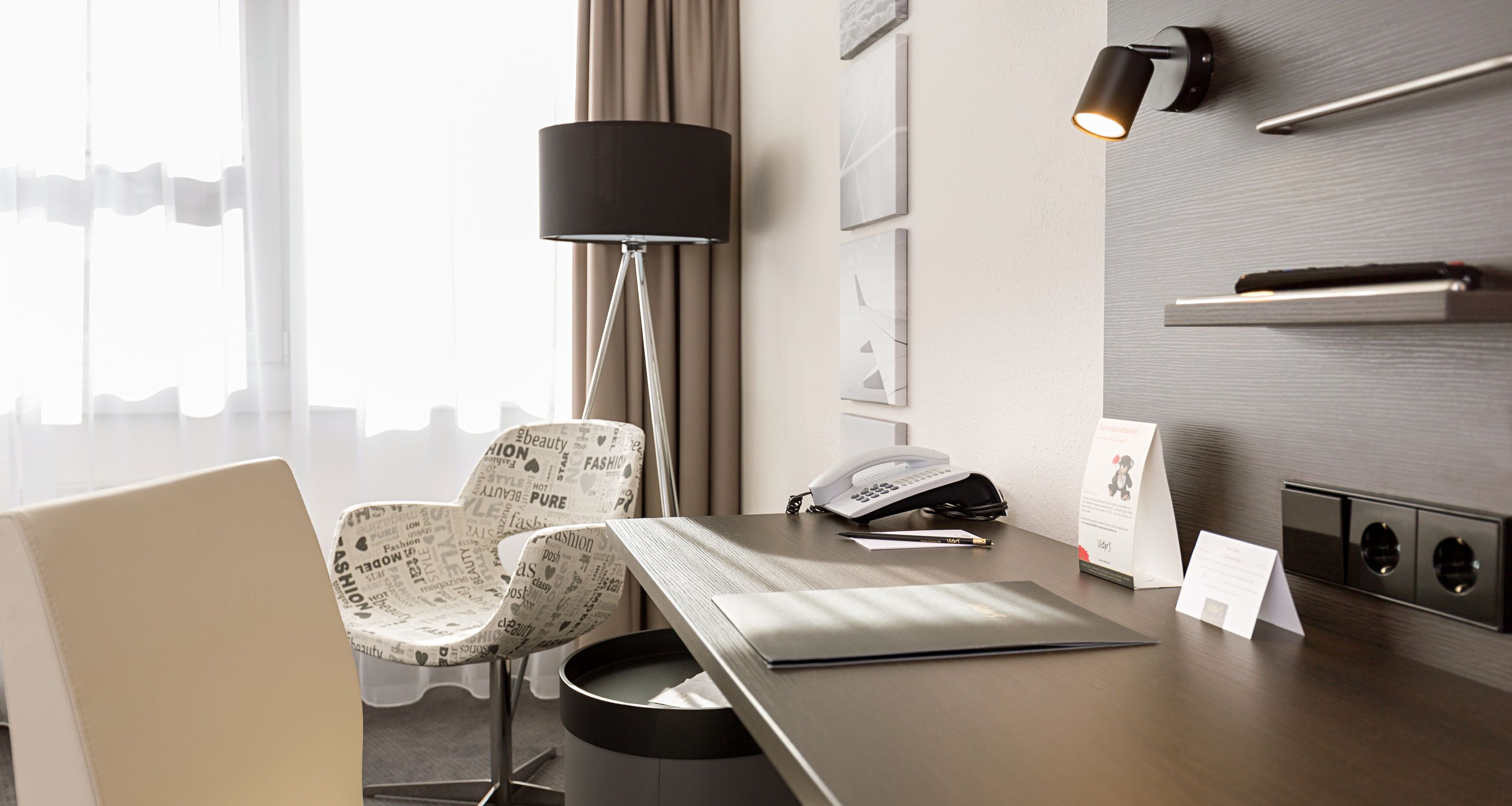Habitación de hotel con escritorio, dos sillones estampados, una lámpara, teléfono y estante de pared.