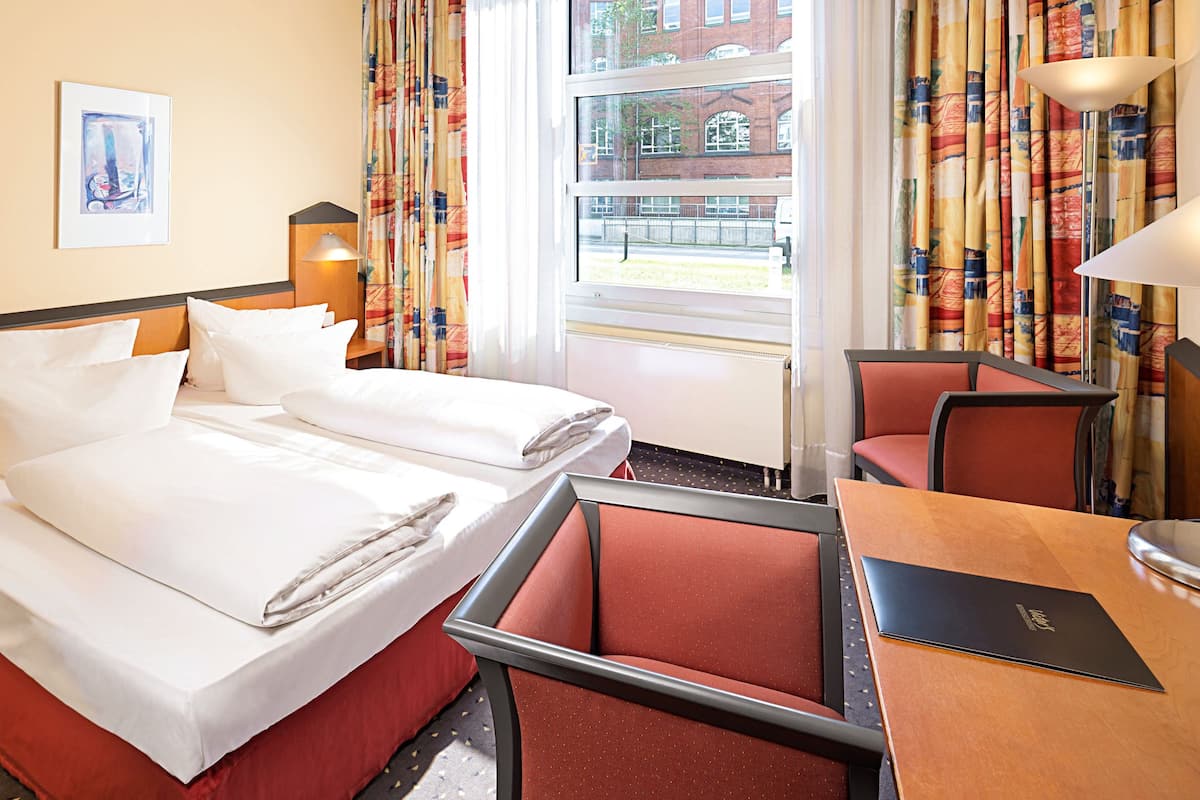 Habitación de hotel con dos camas, un escritorio y una silla. Tiene una ventana con cortinas, una lámpara y un cuadro en la pared.