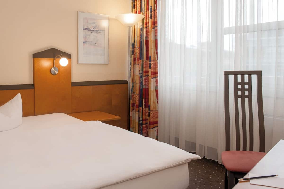 Una habitación de hotel con cama, mesita de noche, lámpara, silla y ventana con cortinas.