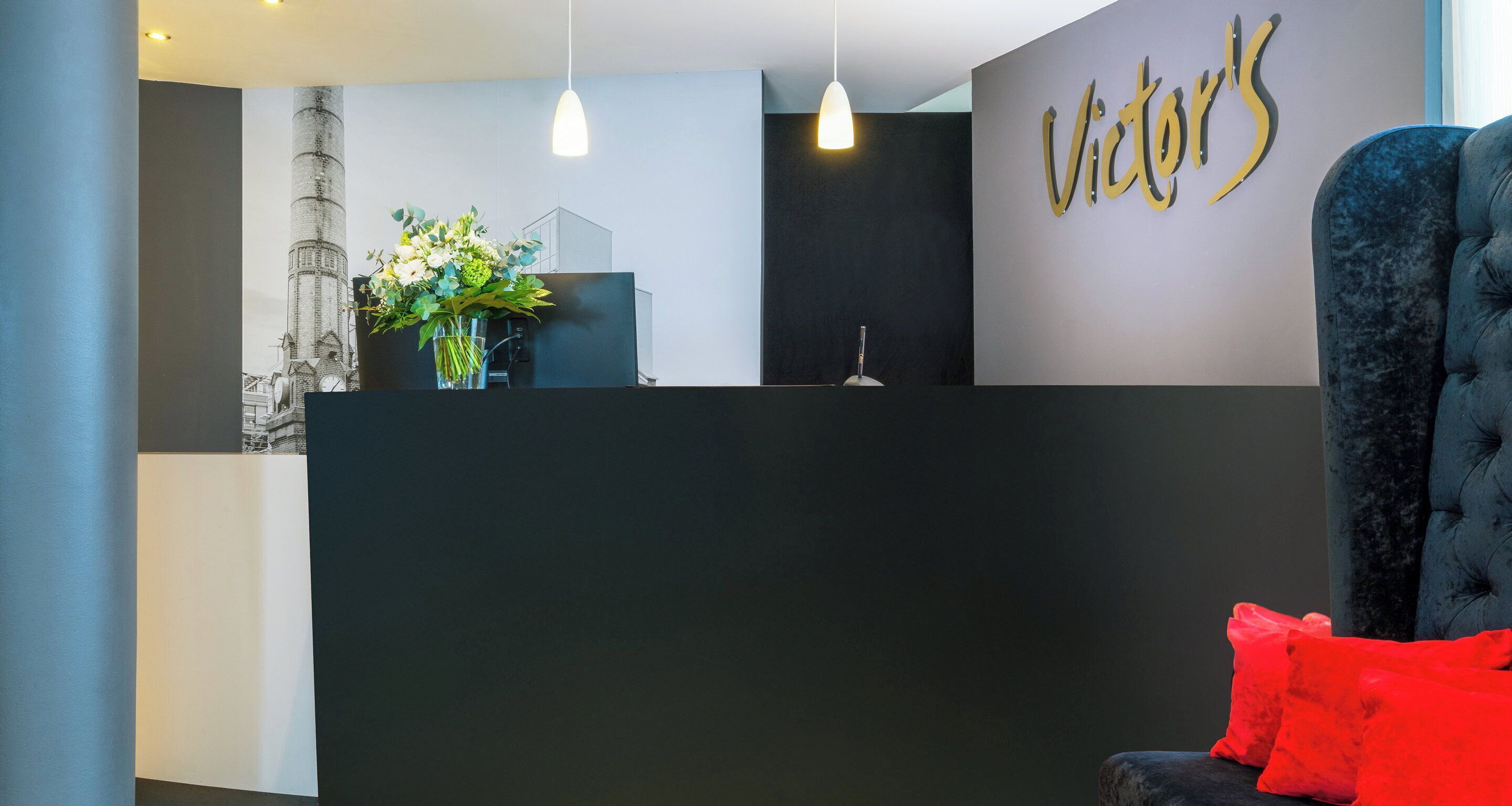 A reception area with a modern design, featuring a black reception desk, a large wall-mounted sign, a potted plant, and a plush blue chair with red cushions.