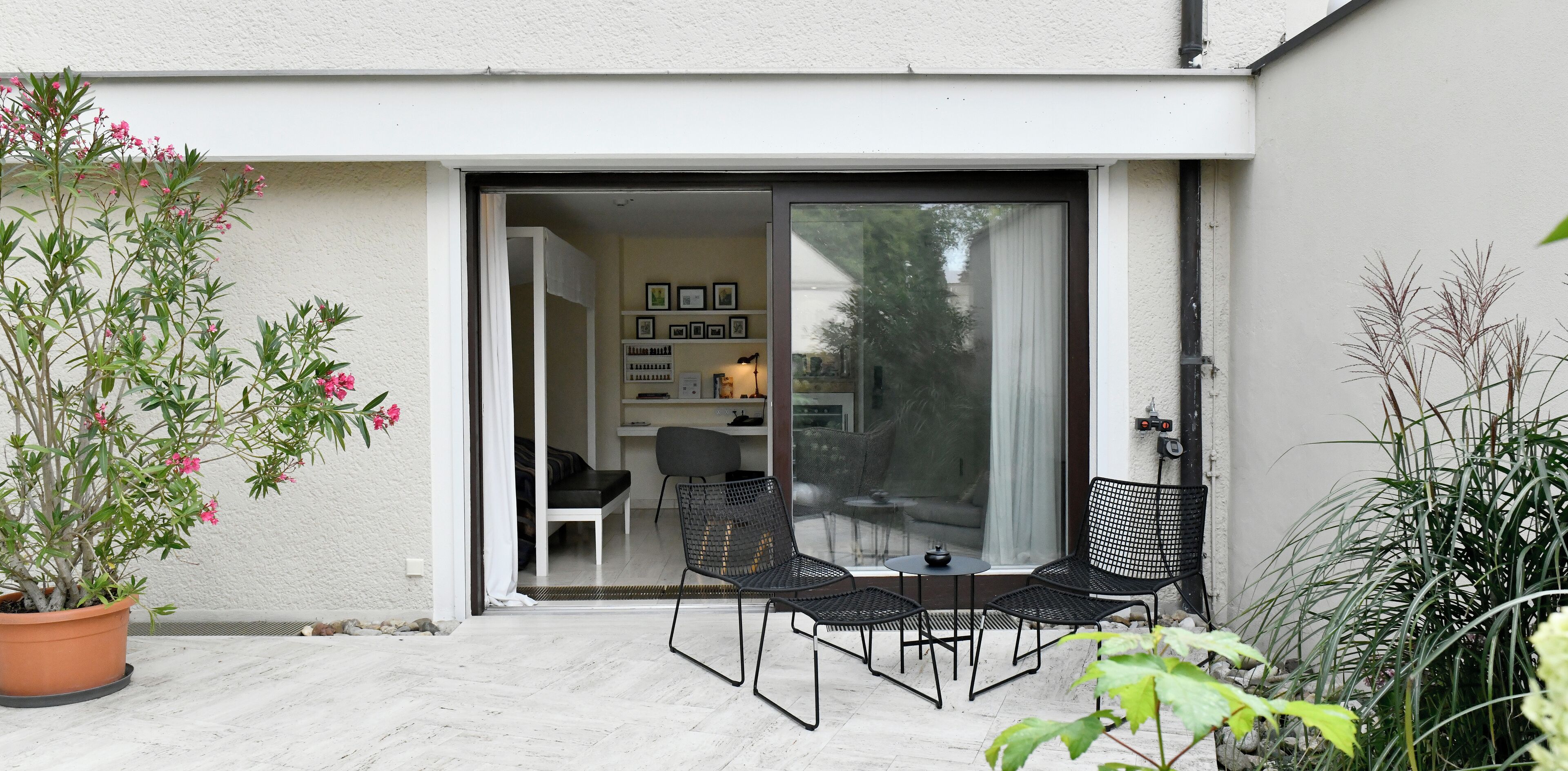 A patio with a table and chairs, a potted plant, and a sliding glass door leading to an interior space with a sofa and a desk.