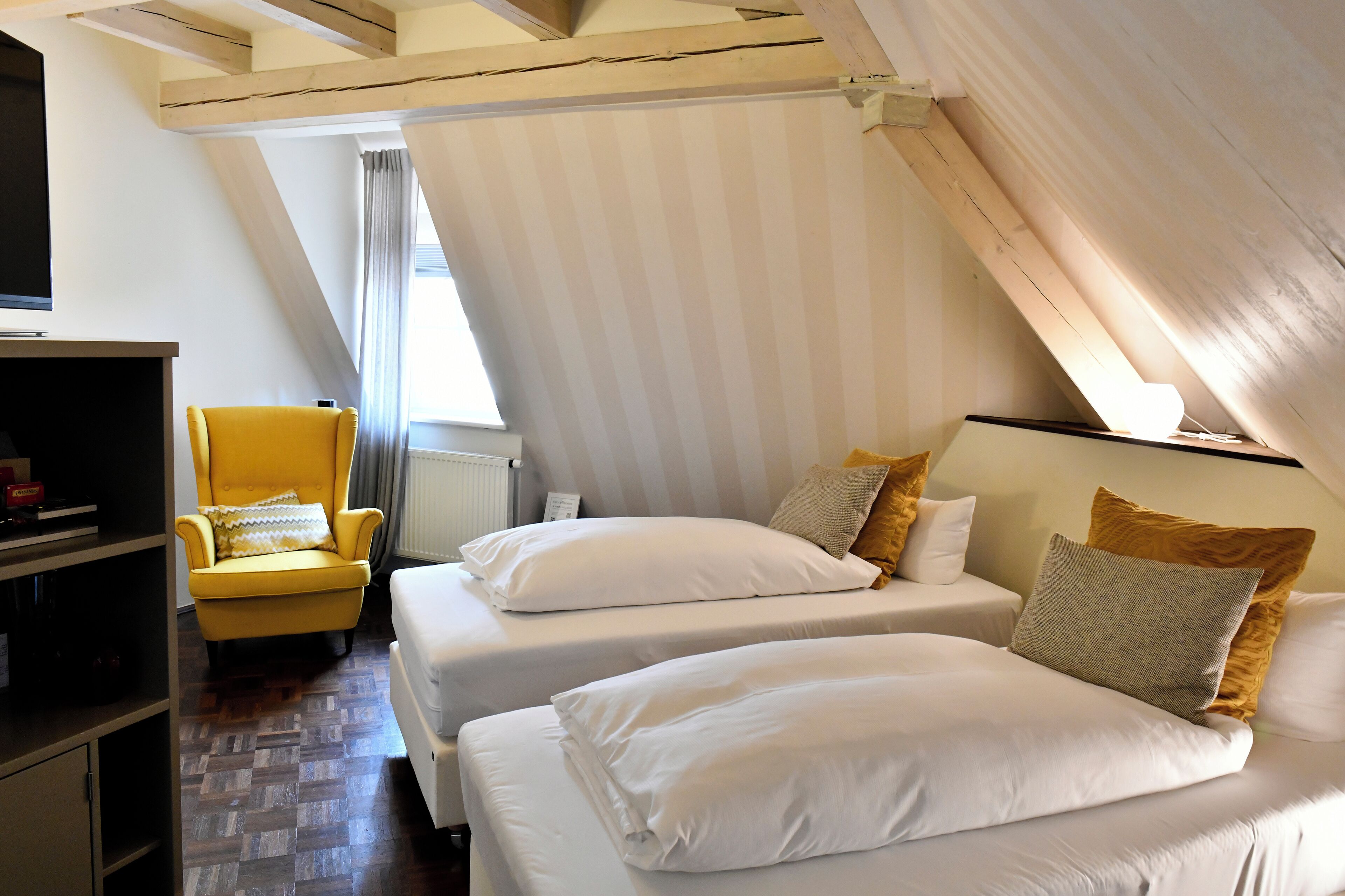 A bedroom with two beds, a yellow armchair, and a wooden ceiling structure.