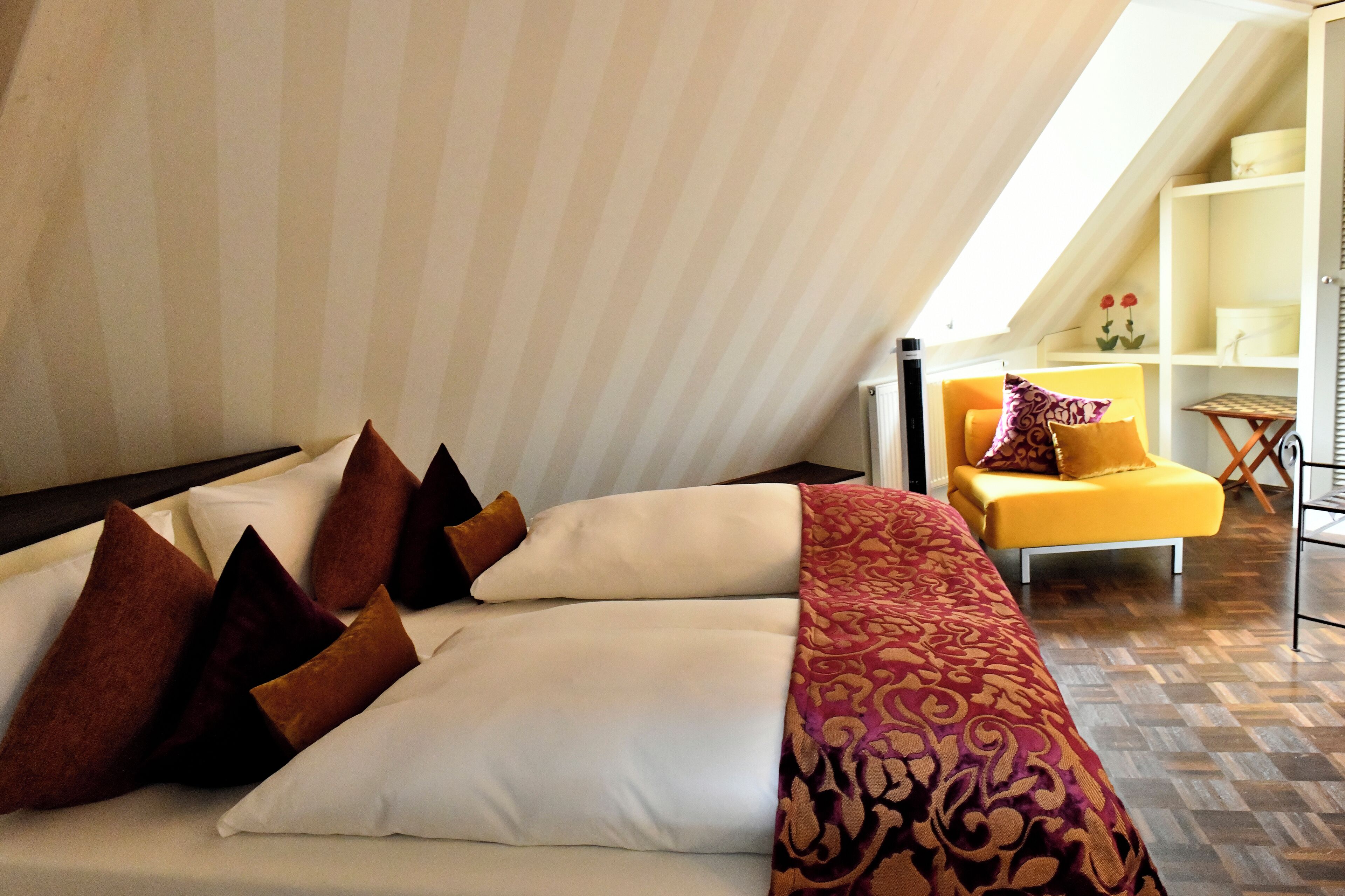 A bedroom with a bed, pillows, a blanket, a yellow chair, and a shelf with decorative items.