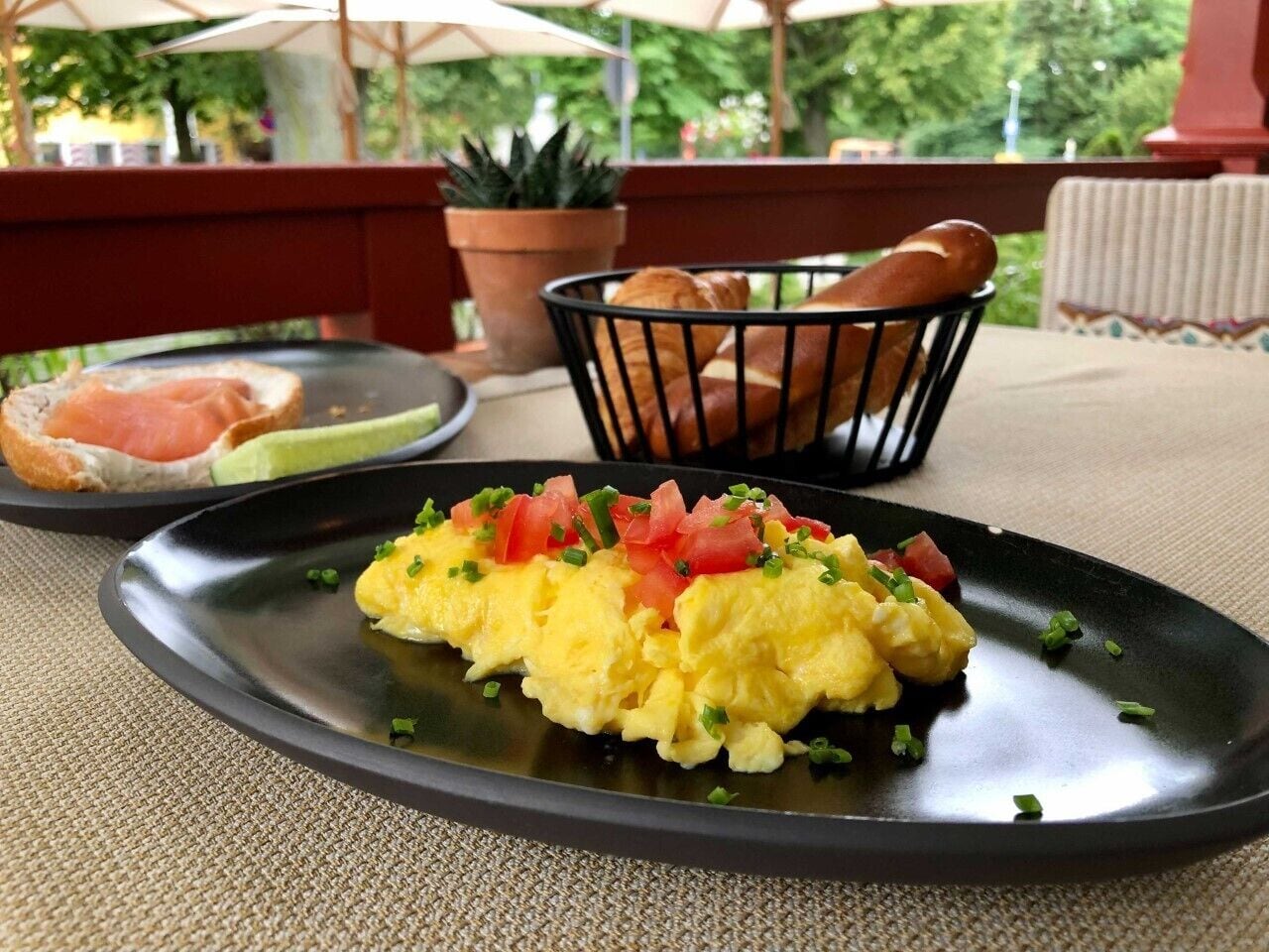 A breakfast plate with scrambled eggs, tomatoes, and chives on a black plate, with bread and a small salad on the side.