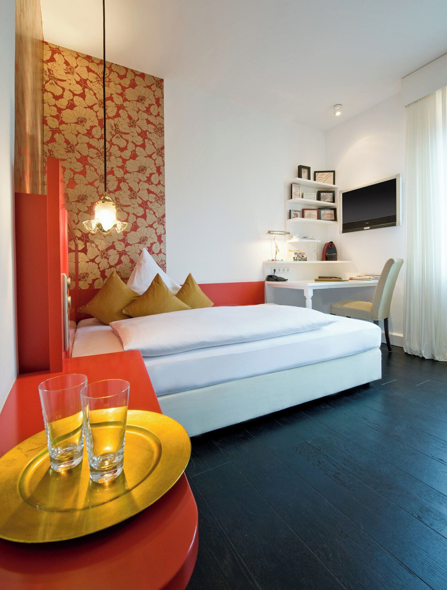 Una habitación de hotel moderna con cama, escritorio y silla. Hay un televisor montado en la pared, una lámpara de escritorio y un pequeño estante con objetos decorativos.