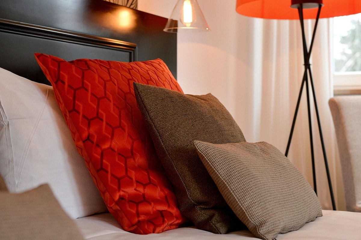 Un sofá con dos almohadones, uno rojo y otro marrón, en una habitación con una lámpara y una ventana.