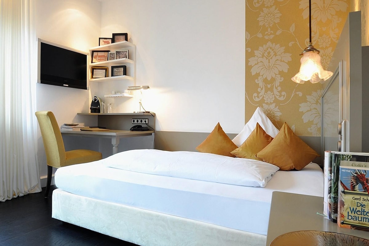 Una habitación de hotel con cama, mesita de noche, escritorio, silla, televisor y estantería.