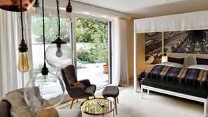 Quarto moderno com uma cama grande, uma cadeira, uma mesa de centro redonda e vista para o jardim através de uma porta deslizante.