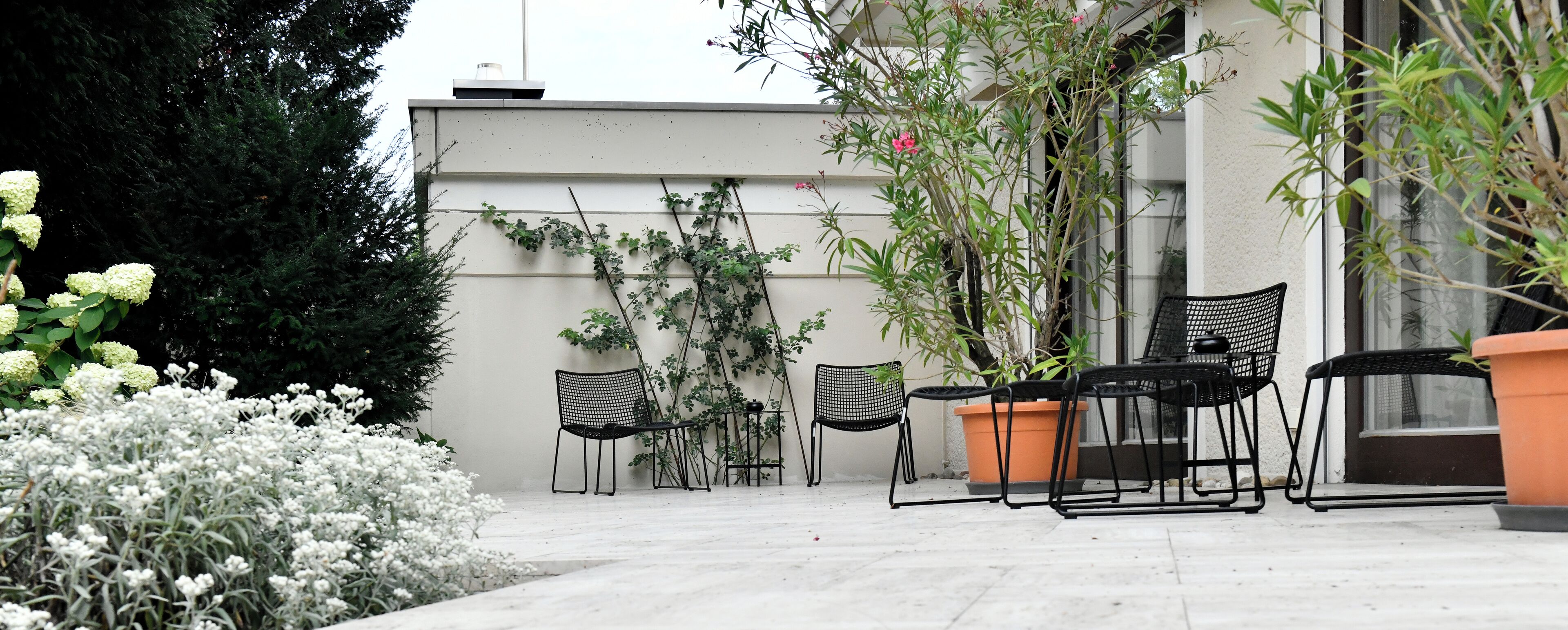 An outdoor patio area with a seating arrangement of two chairs and a table, surrounded by potted plants and flowering shrubs.