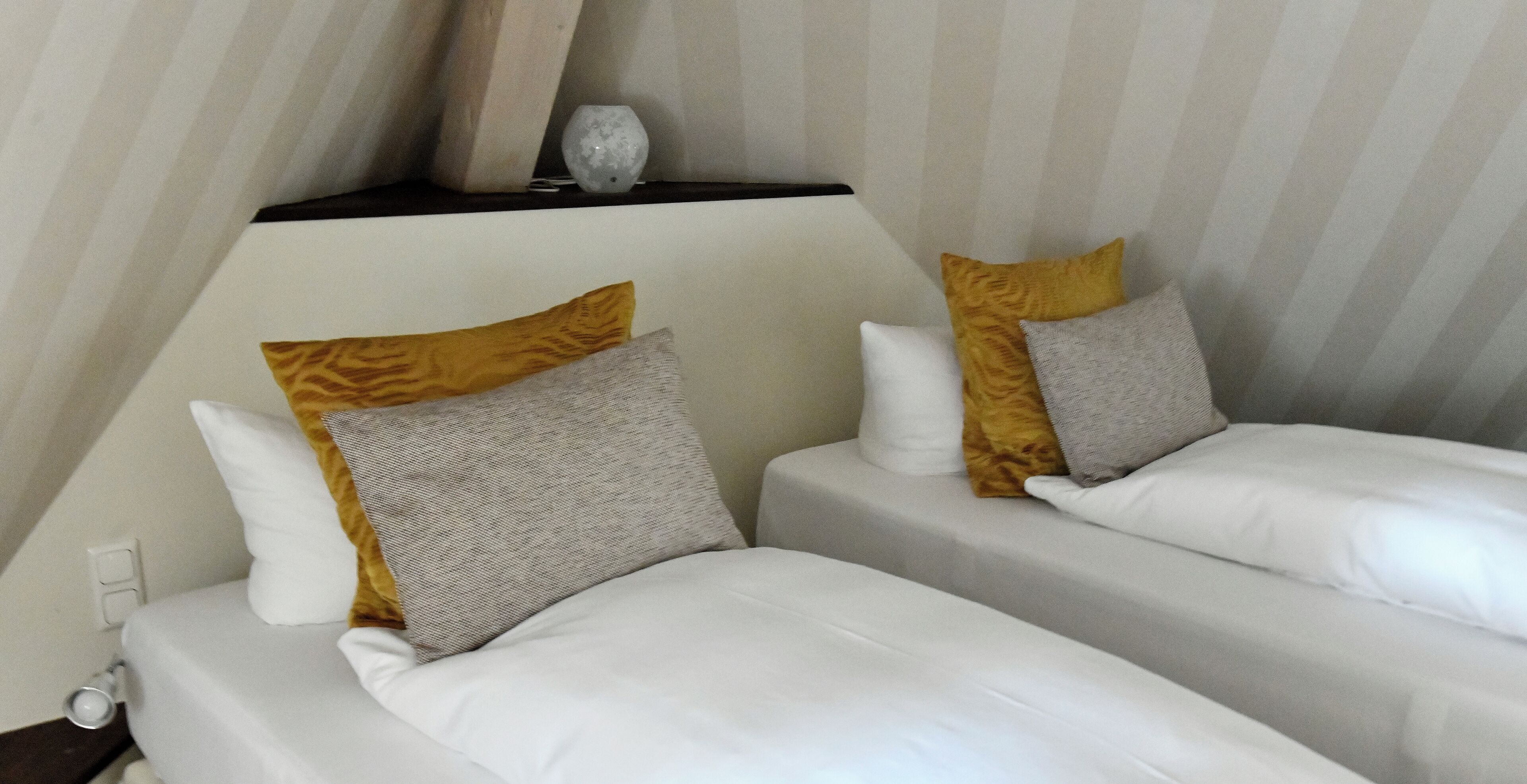 Two single beds with white and patterned bedding, a small table with a decorative item, and a striped wall.