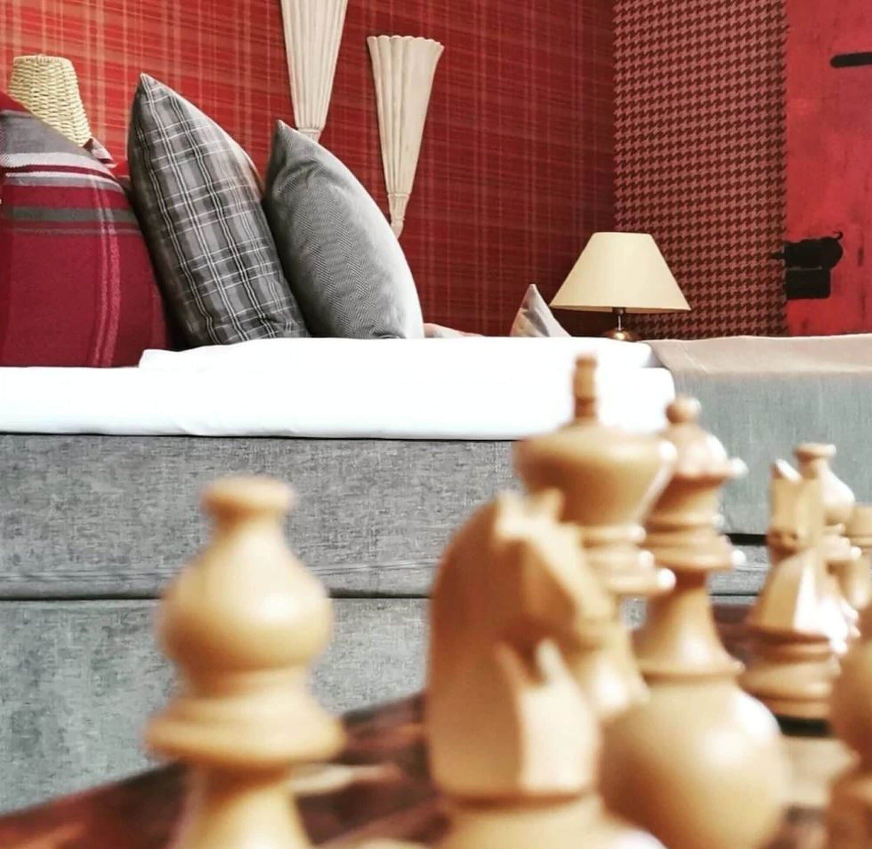 A chessboard with wooden pieces in focus, a sofa with a plaid blanket and pillows, and a lamp on a table.