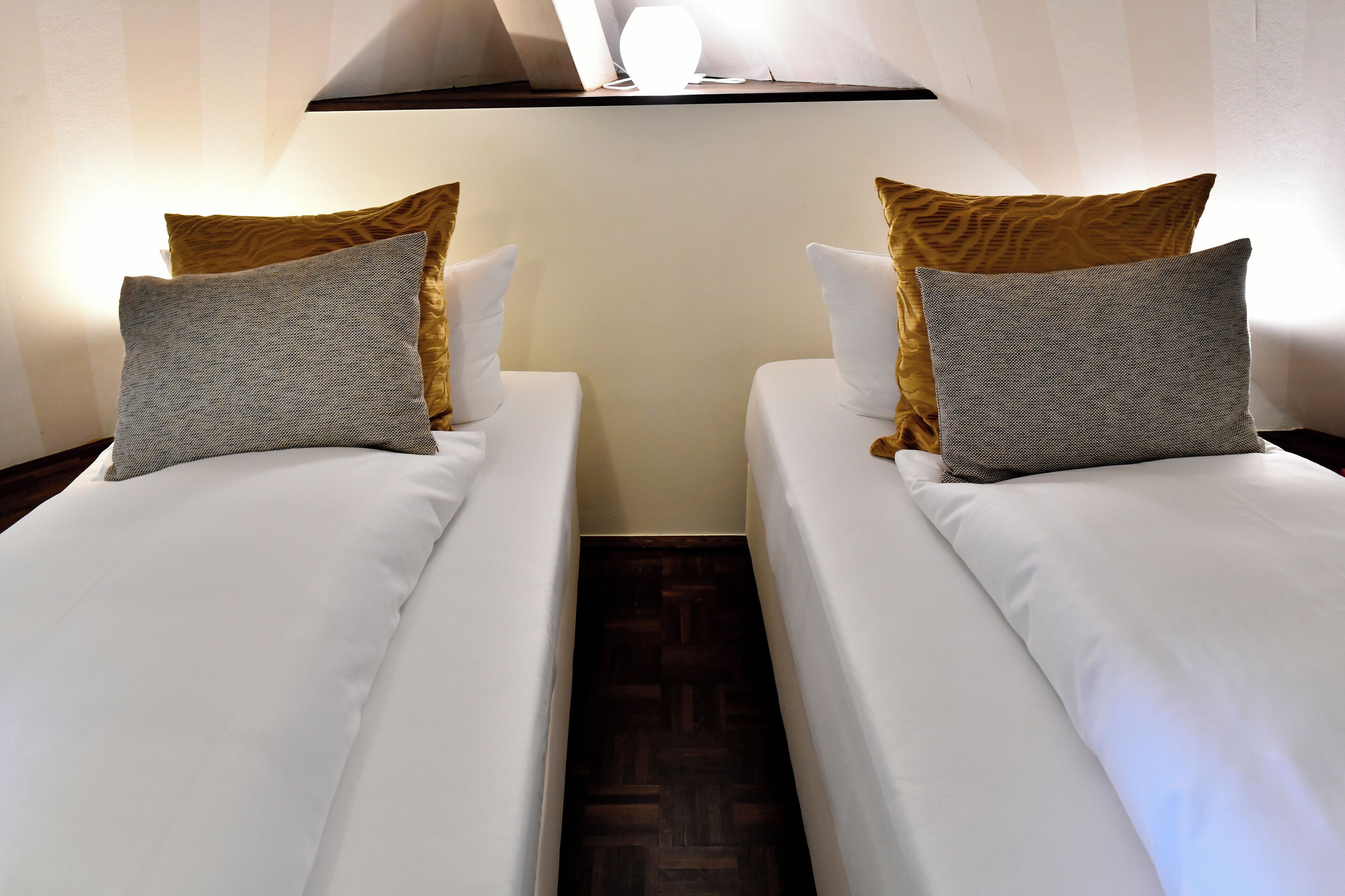 Deux lits simples avec du linge de lit blanc et des oreillers gris, un plancher en bois et un mur blanc avec un luminaire au-dessus.
