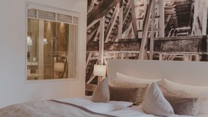 Quarto com cama grande, janela com vista para uma estrutura de madeira e luminária fixada na parede.