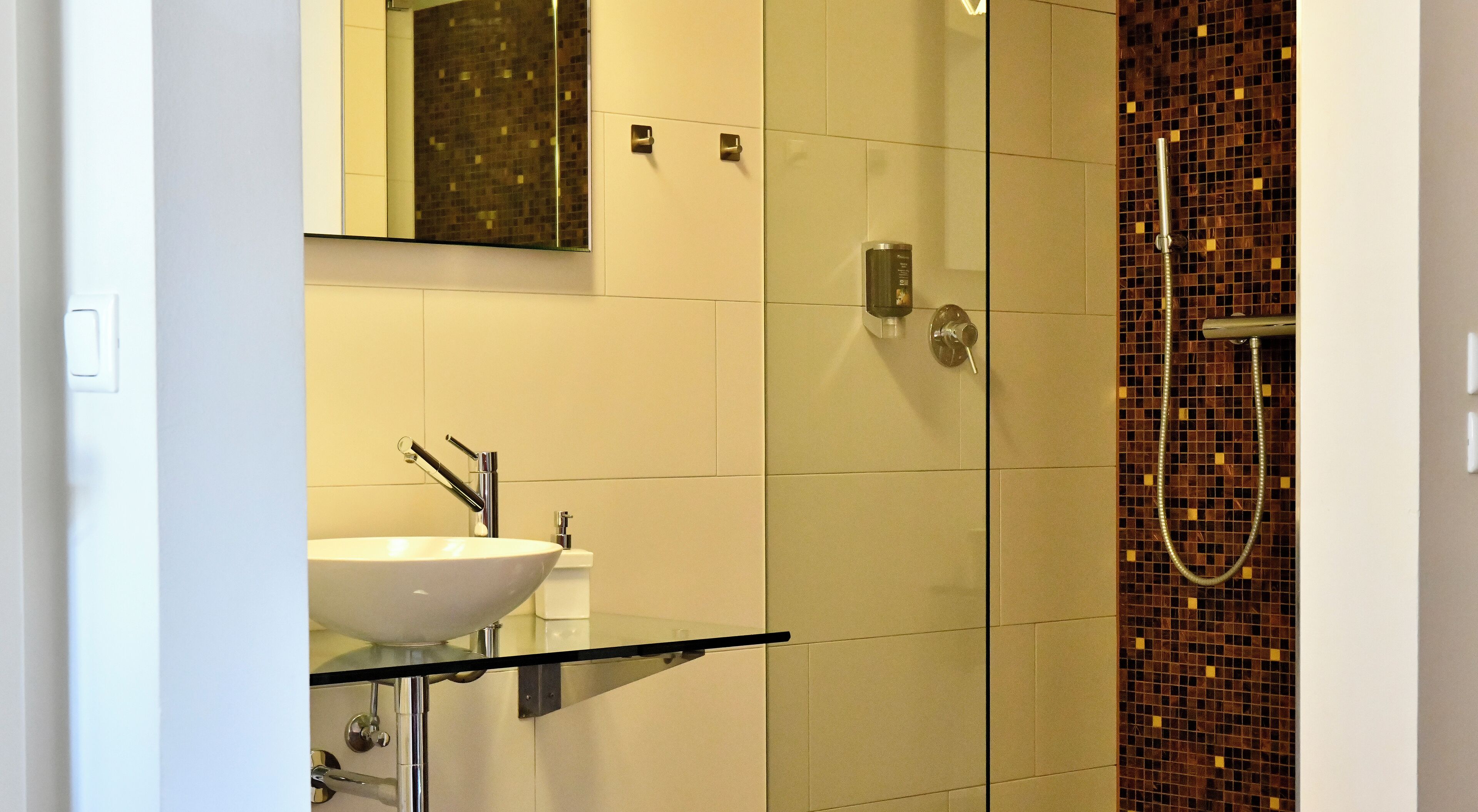 Un baño moderno con un lavamanos con cubierta de vidrio, una ducha con pared de mosaicos y un espejo sobre el lavamanos.