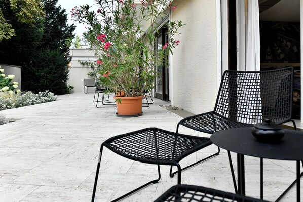 An outdoor patio with two black metal chairs and a table, a potted plant with pink flowers, and a white wall in the background.
