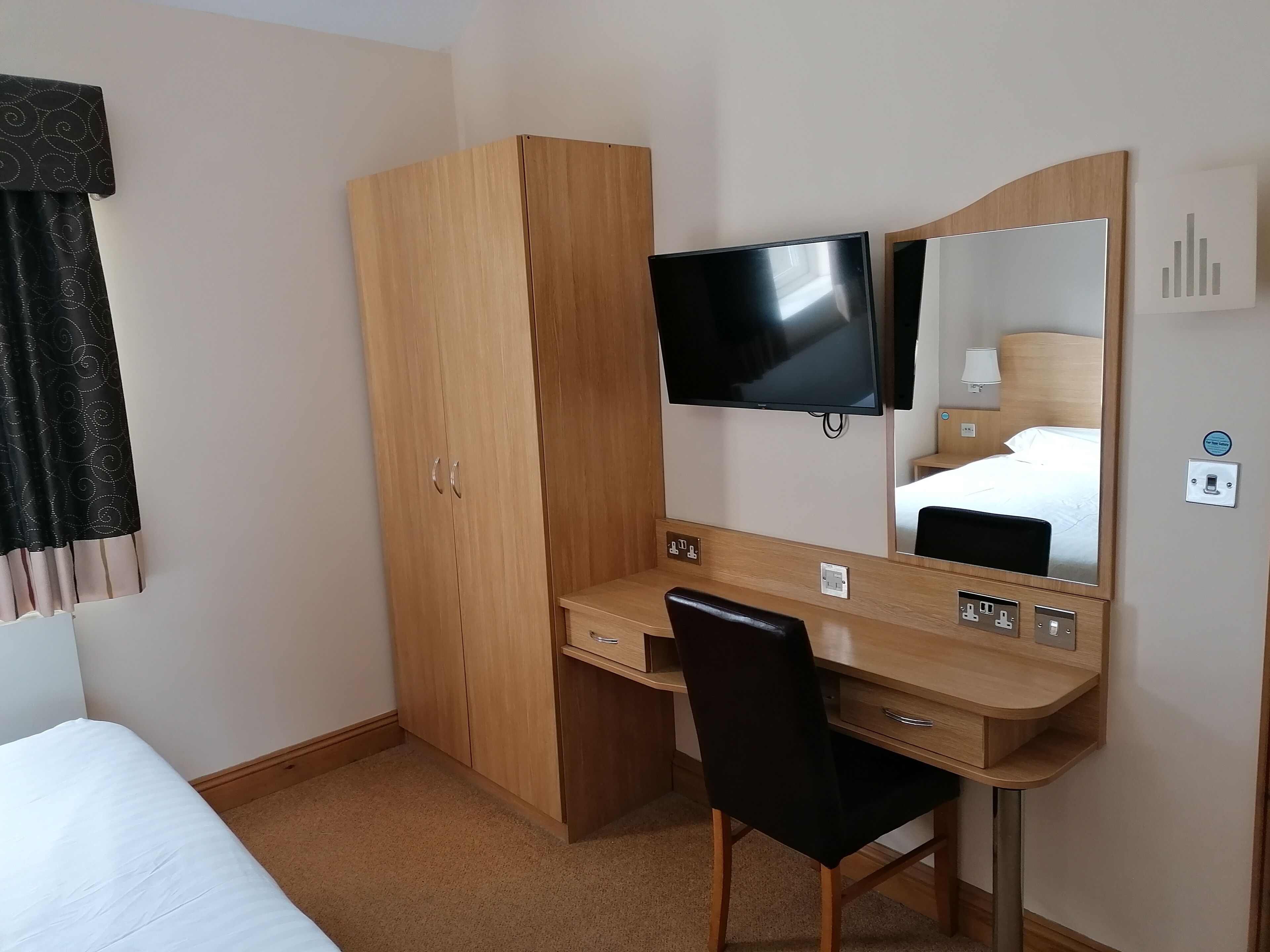 A hotel room with a wooden desk, a black chair, a flat-screen TV, a large mirror, and a bed with white bedding.