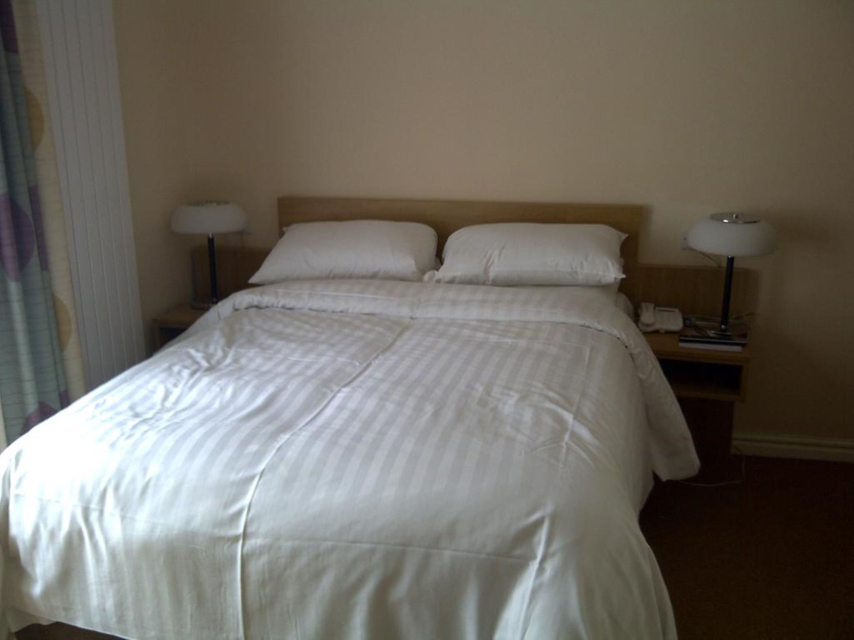 A neatly made bed with white linens, two bedside lamps, and a nightstand with a phone and a lamp.
