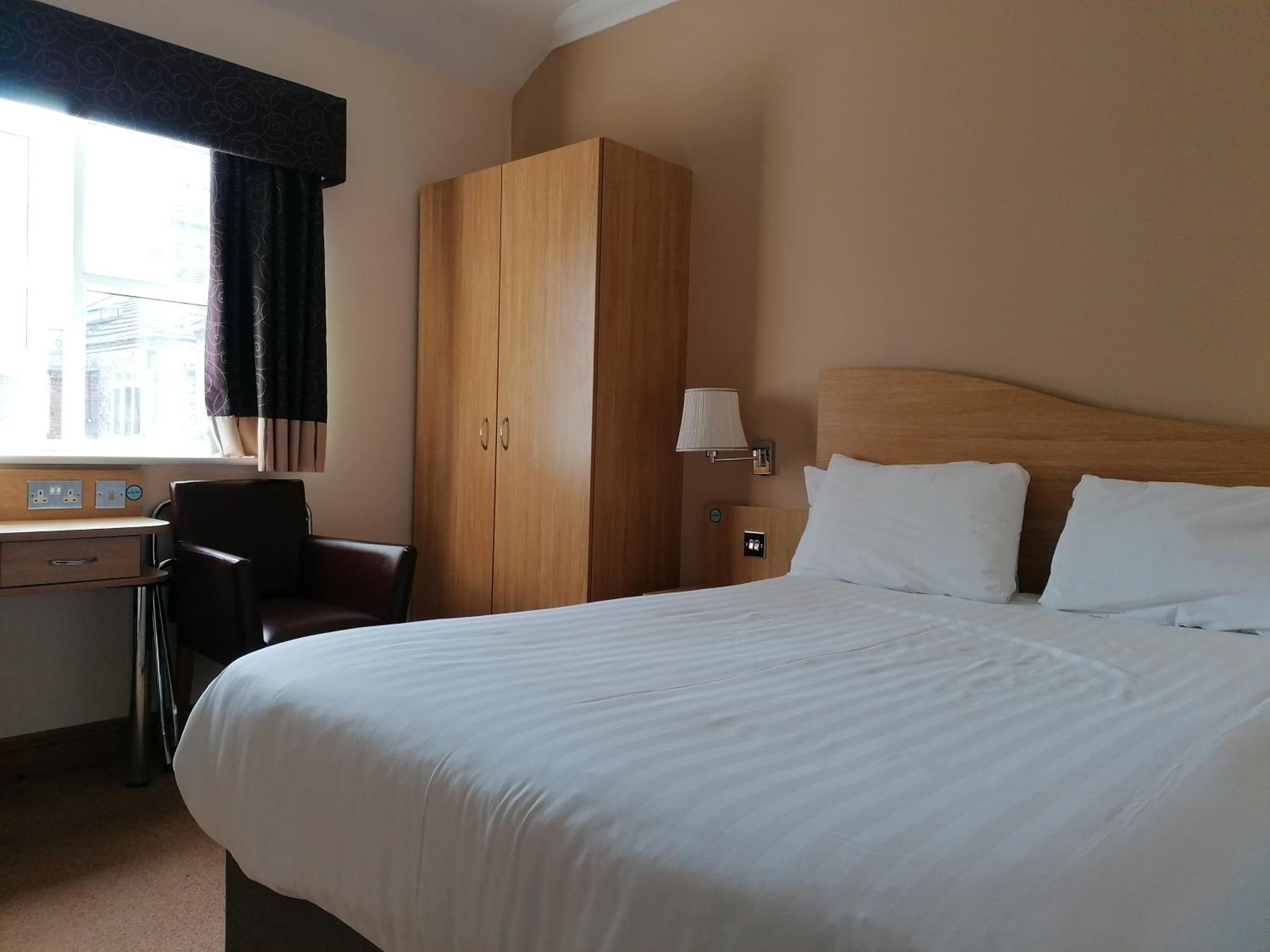 A hotel room with a bed, a desk, a chair, a wardrobe, and a window with curtains.