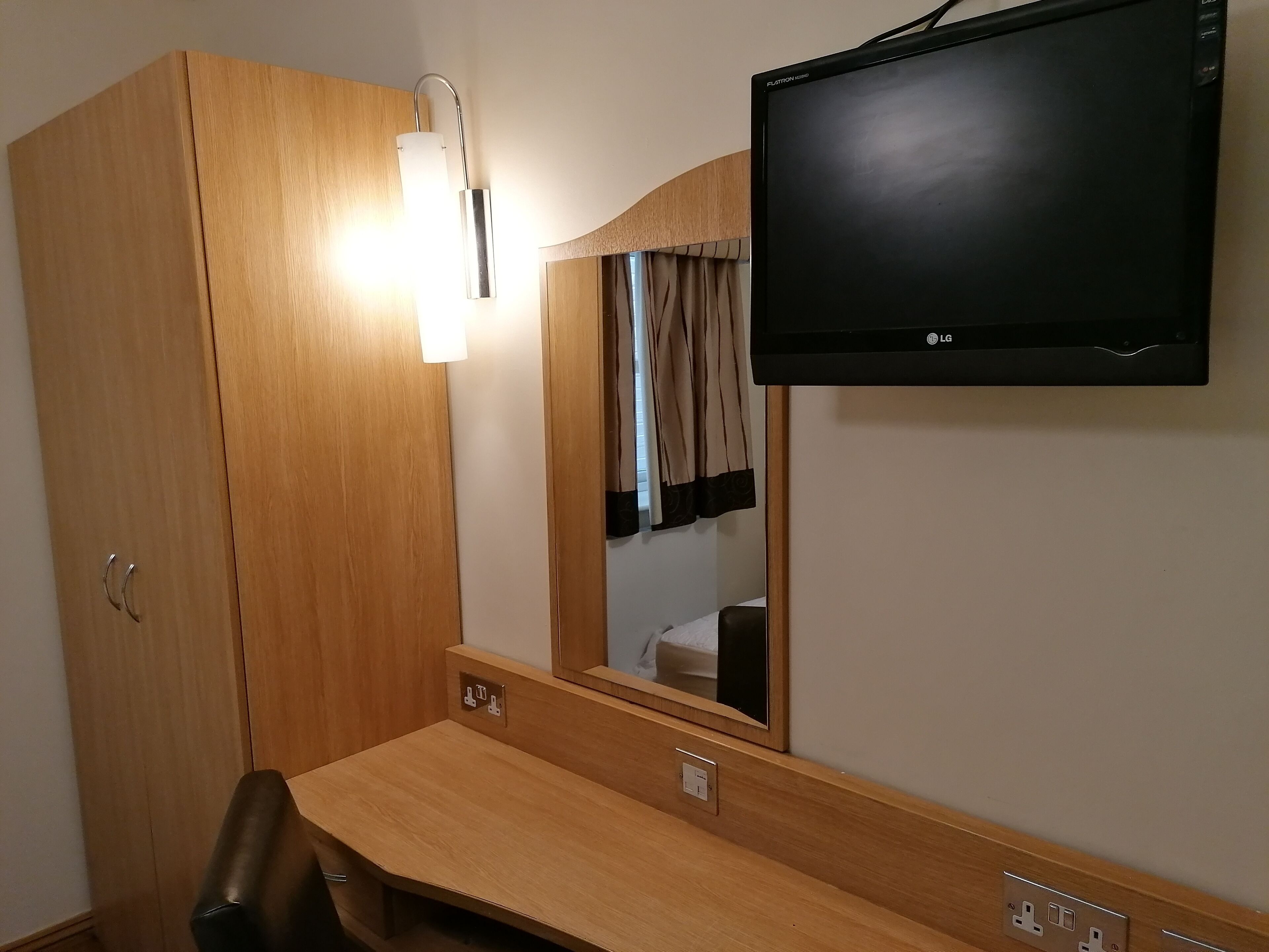 A hotel room with a wooden wardrobe, a bedside table, a mirror, and a flat-screen TV mounted on the wall.