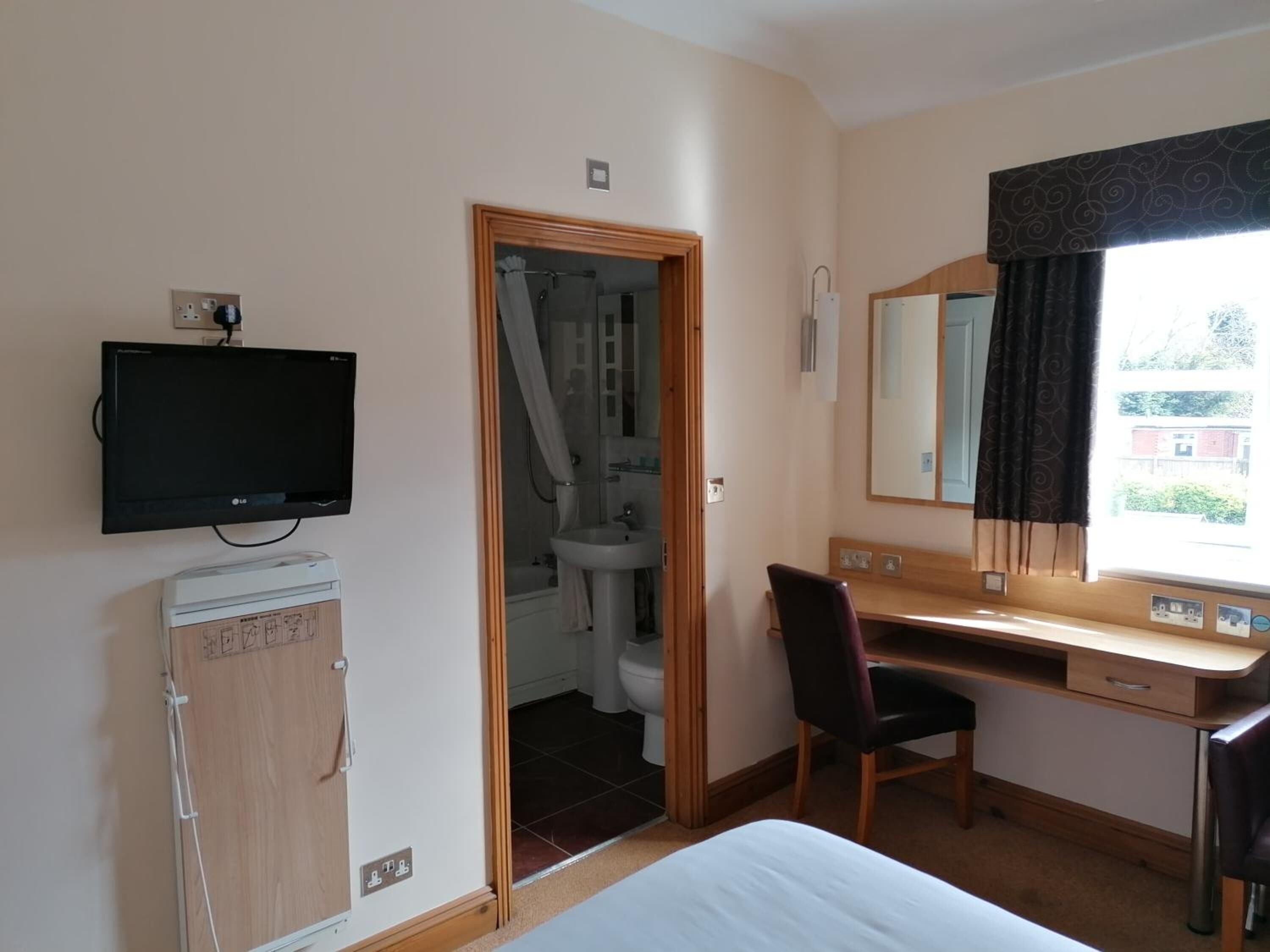 A hotel room with a bed, a desk with a chair, a television, a bathroom with a shower, and a window with curtains.