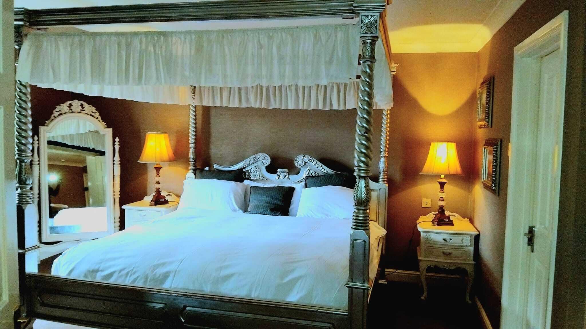 A four-poster bed with a canopy, two bedside tables with lamps, a mirror, and a door to the right.