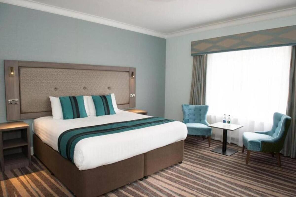 A hotel room with a large bed, a bedside table, a chair, a small table, and a window with curtains.