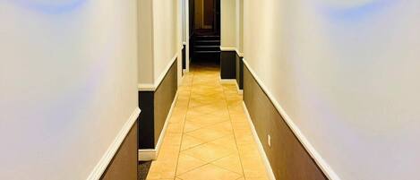 Pasillo de hotel con piso de baldosas, paredes blancas y una sección inferior de color oscuro.