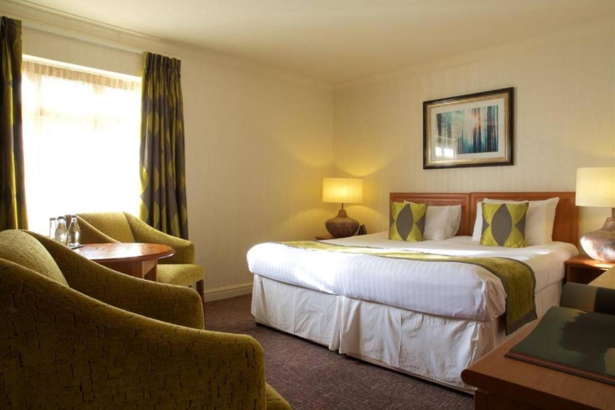 A hotel room with a bed, two chairs, a table, a lamp, and a painting on the wall.