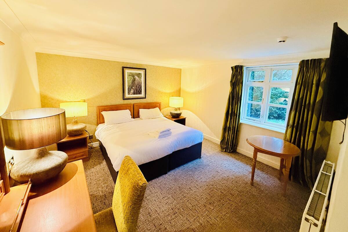 A hotel room with a bed, two bedside tables, a chair, a small table, and a window with curtains.