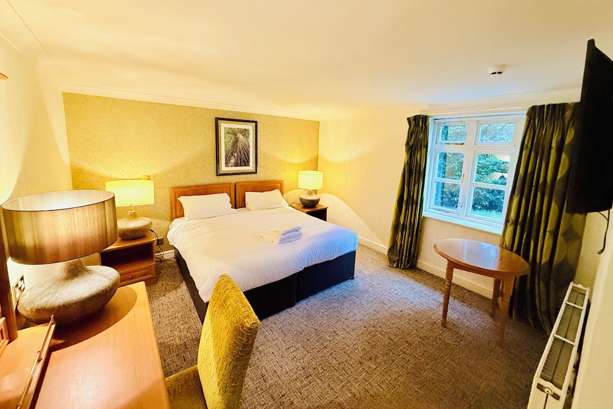 A hotel room with a bed, two bedside tables, a chair, a small table, and a window with curtains.