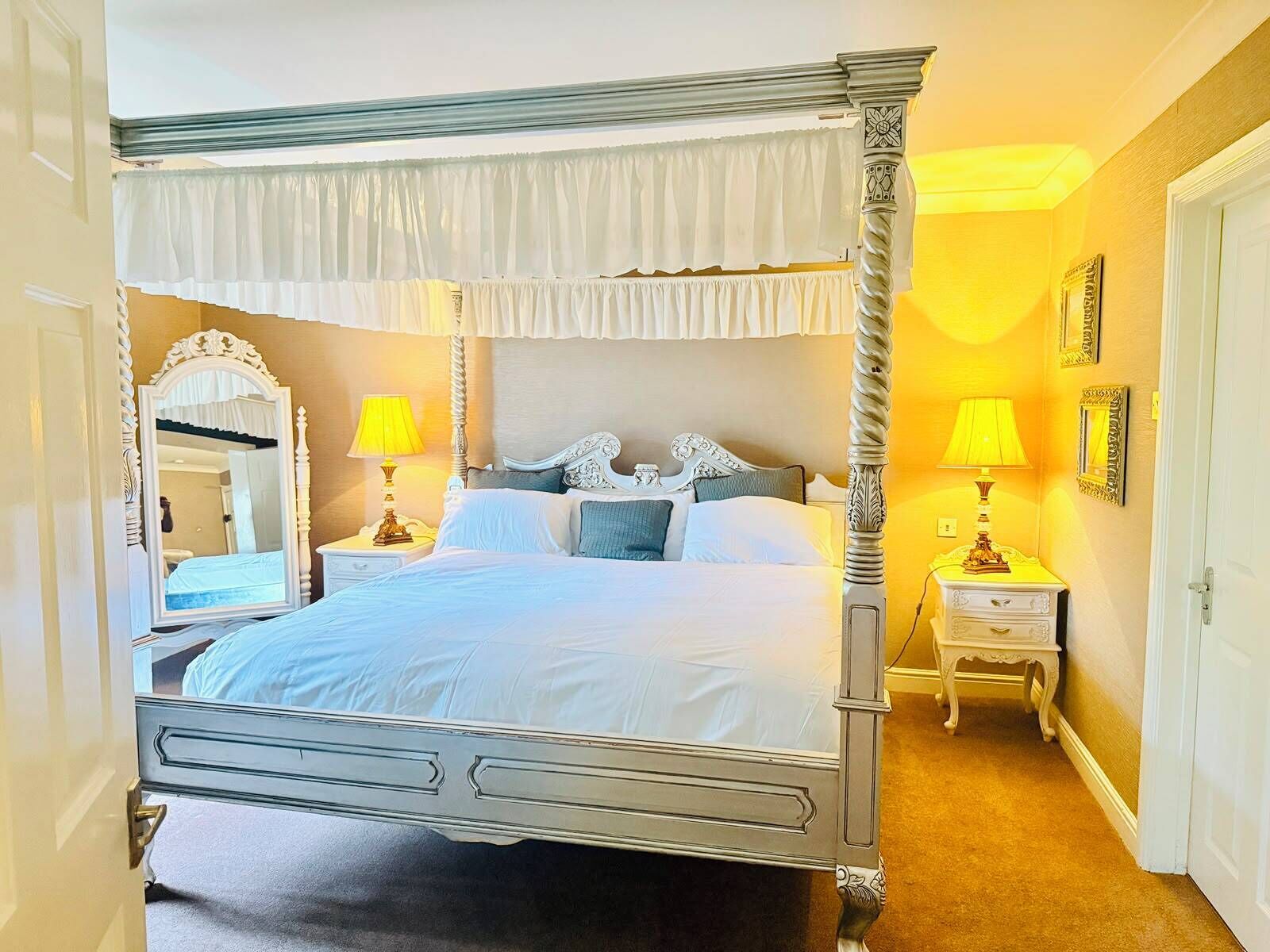 Una cama con dosel, dos mesitas de noche con lámparas, un espejo y una puerta a la izquierda.