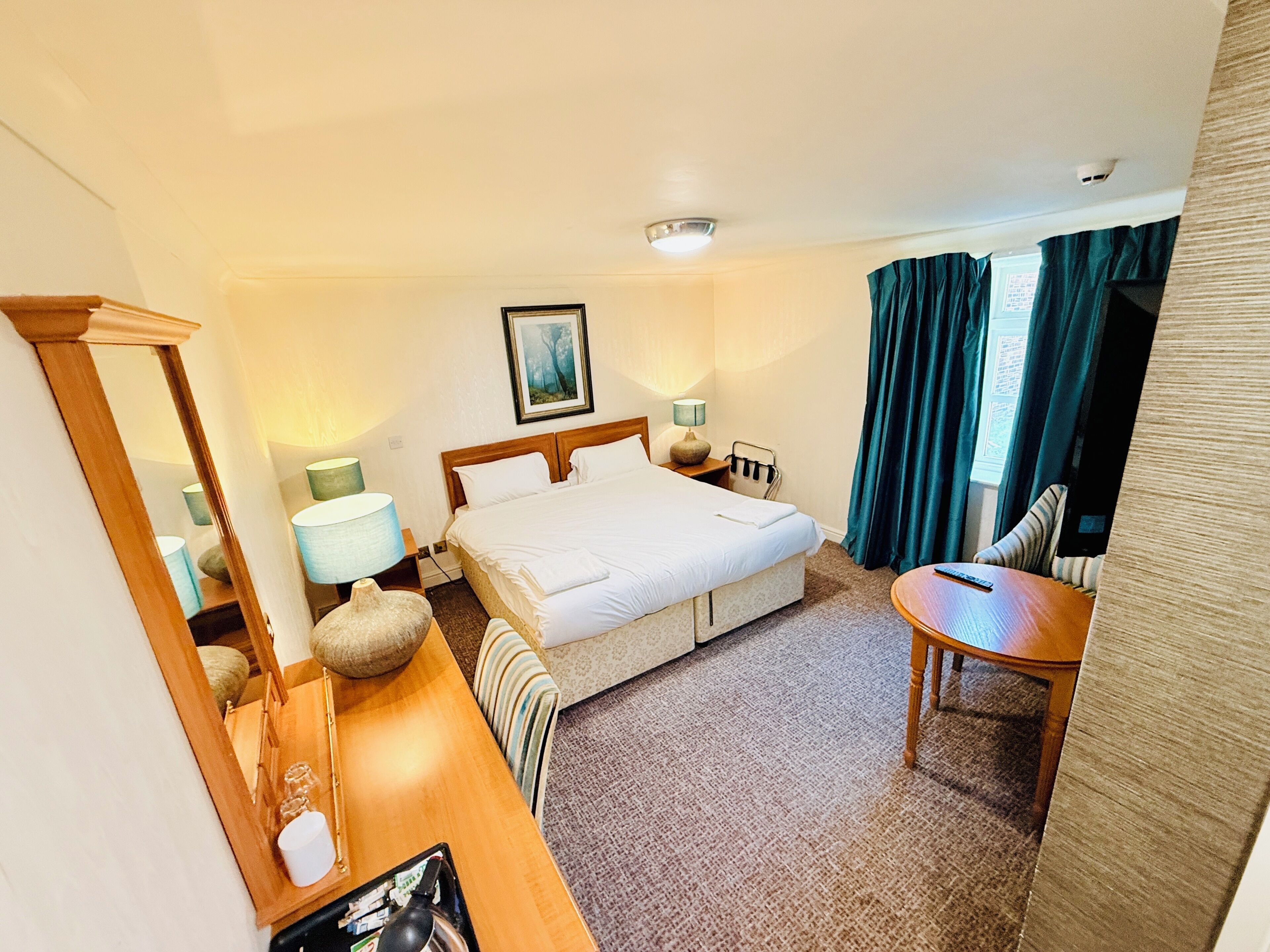 A hotel room with a bed, bedside tables, a desk, a chair, a lamp, a picture on the wall, and a window with curtains.