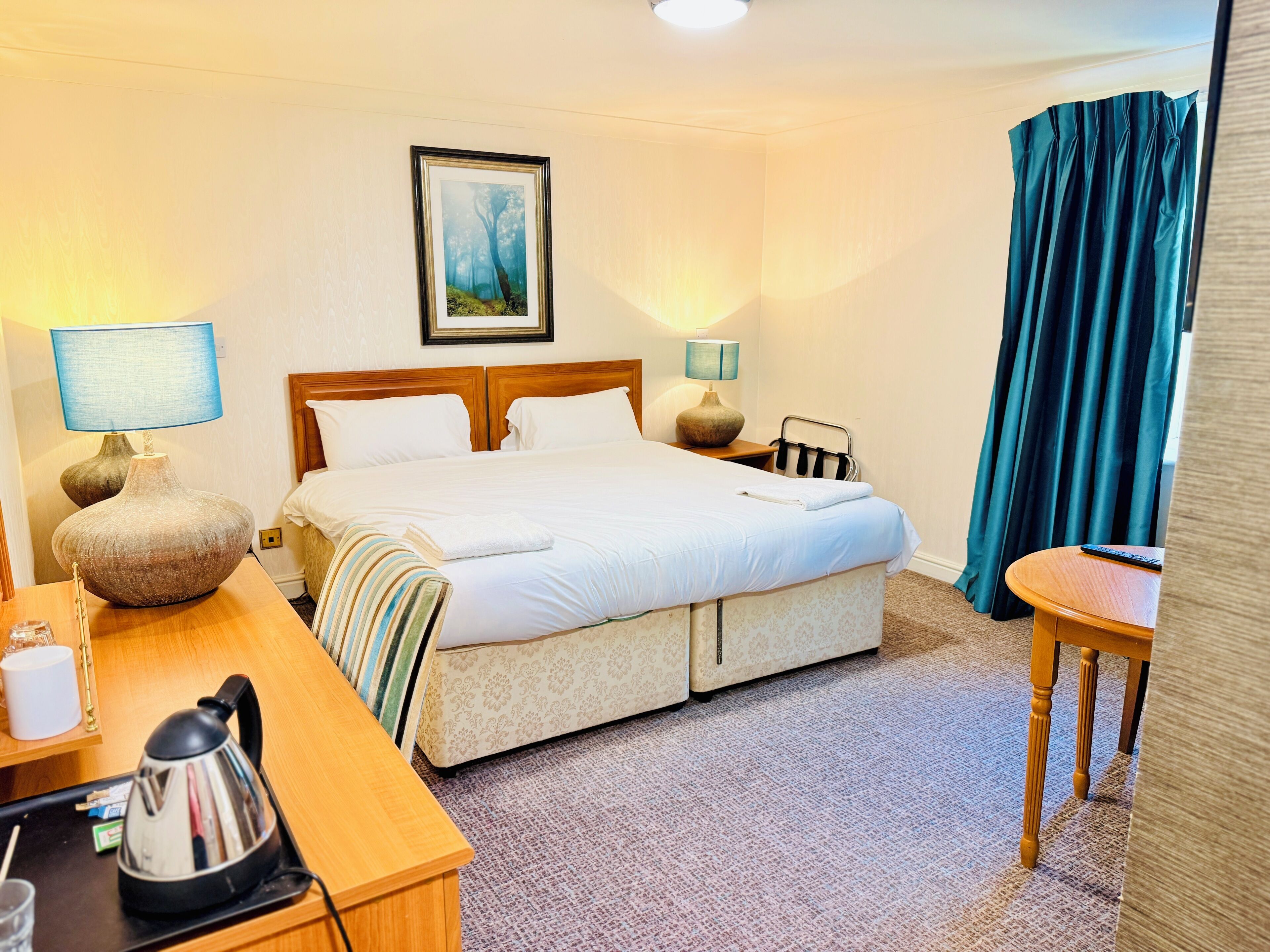 A hotel room with a bed, bedside lamps, a picture on the wall, a desk with a kettle, and a chair.