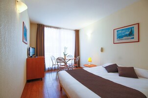 Habitación de hotel con una cama, un escritorio con computadora, una silla, una mesa con una planta y una lámpara.