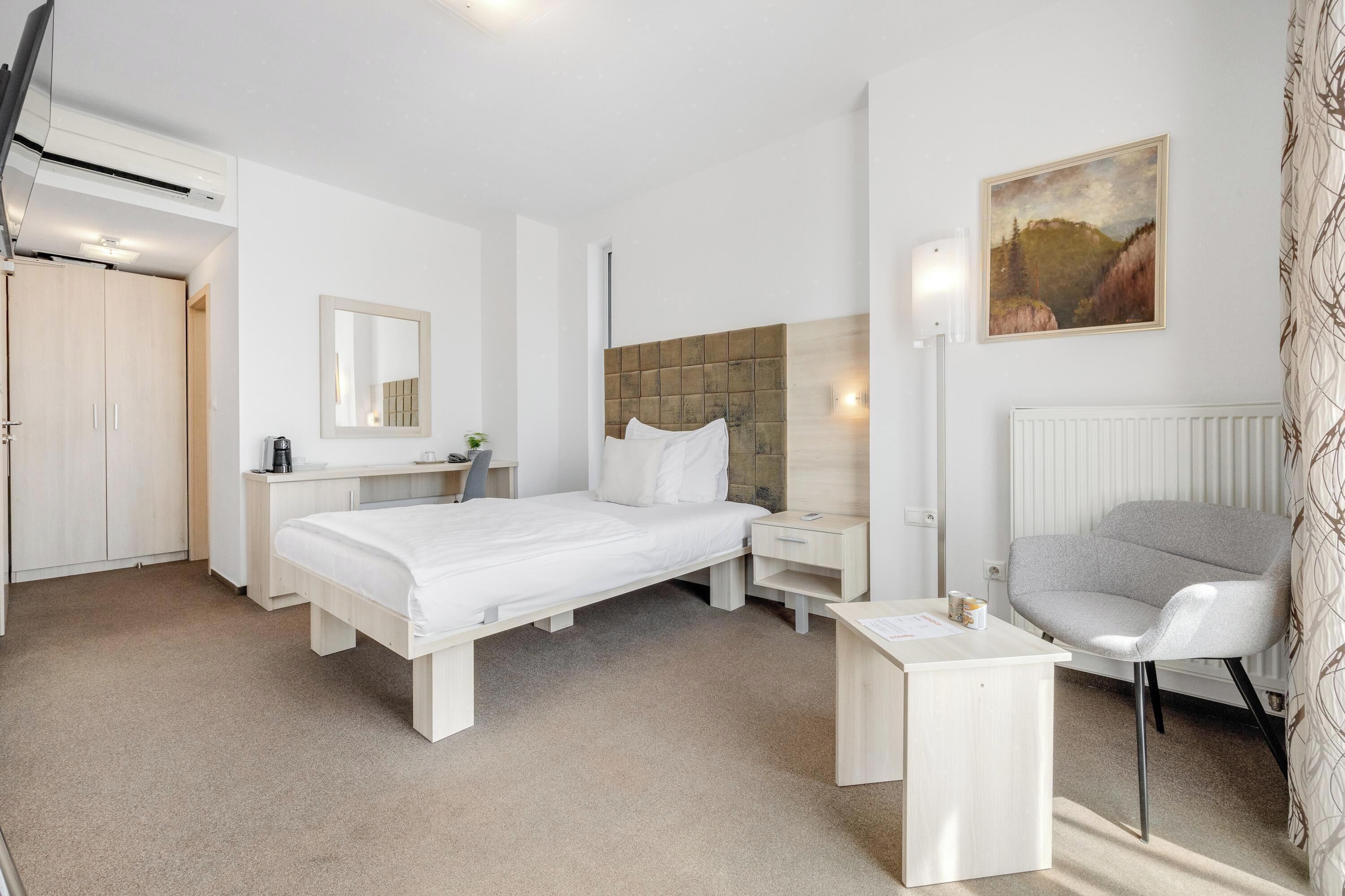 A hotel room with a bed, bedside table, chair, desk, and a painting on the wall.