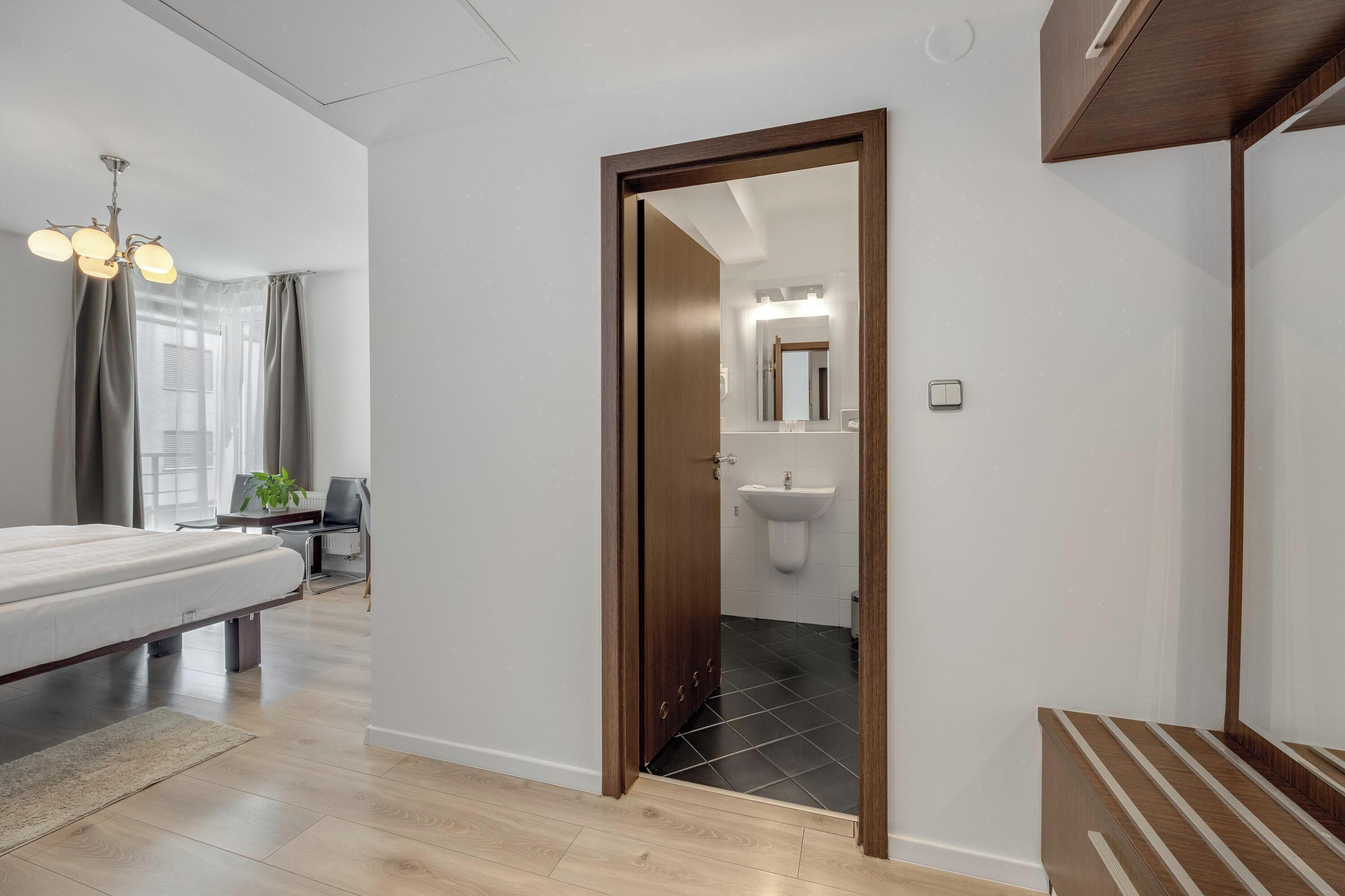Un dormitorio moderno con cama, mesita de noche y escritorio con silla. Se ve un baño con lavamanos y espejo a través de una puerta abierta.
