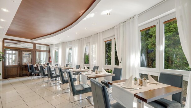 A modern hotel dining area with multiple tables and chairs, large windows with curtains, and a visible bar area. - Hotel ANTARES (Bratislava)
