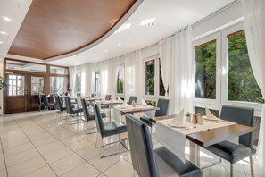 A modern hotel dining area with multiple tables and chairs, large windows with curtains, and a visible bar area. - Hotel ANTARES (Bratislava)