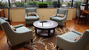 A seating area with three grey armchairs and a central glass-top table, a wooden floor, and a view of parked cars through large windows.