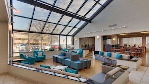 A spacious indoor area with a high ceiling and large windows, featuring multiple seating areas with sofas and chairs, and a visible outdoor view through the windows.