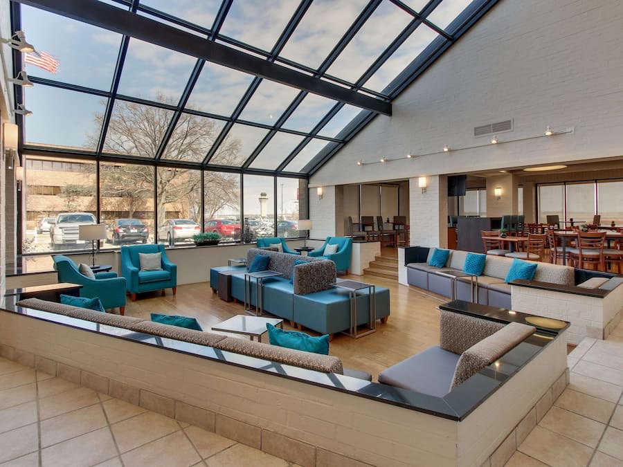 A spacious indoor area with a high ceiling and large windows, featuring multiple seating areas with sofas and chairs, and a visible outdoor view through the windows.