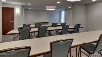 Un salón de conferencias con una mesa larga y varias sillas, equipado con luces en el techo y una ventana con persianas.