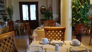Un comedor con mesas dispuestas para cuatro comensales, cada una con platos, tazones, cubiertos y servilletas.  Sillas rodean las mesas, y hay plantas en macetas cerca de ellas.