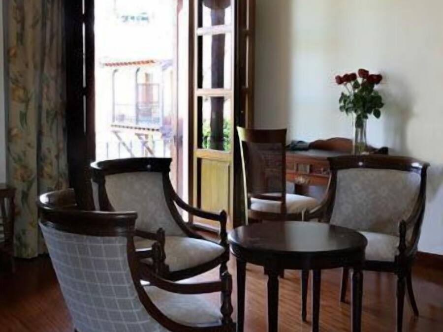 Una habitación con suelo de madera, una mesa de comedor con sillas, una ventana con cortinas y un jarrón con flores sobre una mesa.