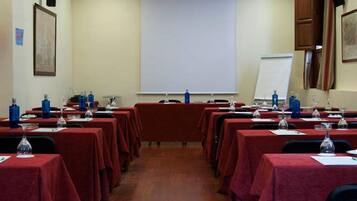 Un salón de conferencias con filas de mesas cubiertas con manteles rojos, cada una con un vaso y una botella de agua. Hay una pantalla de proyector al frente y una pizarra lateral.