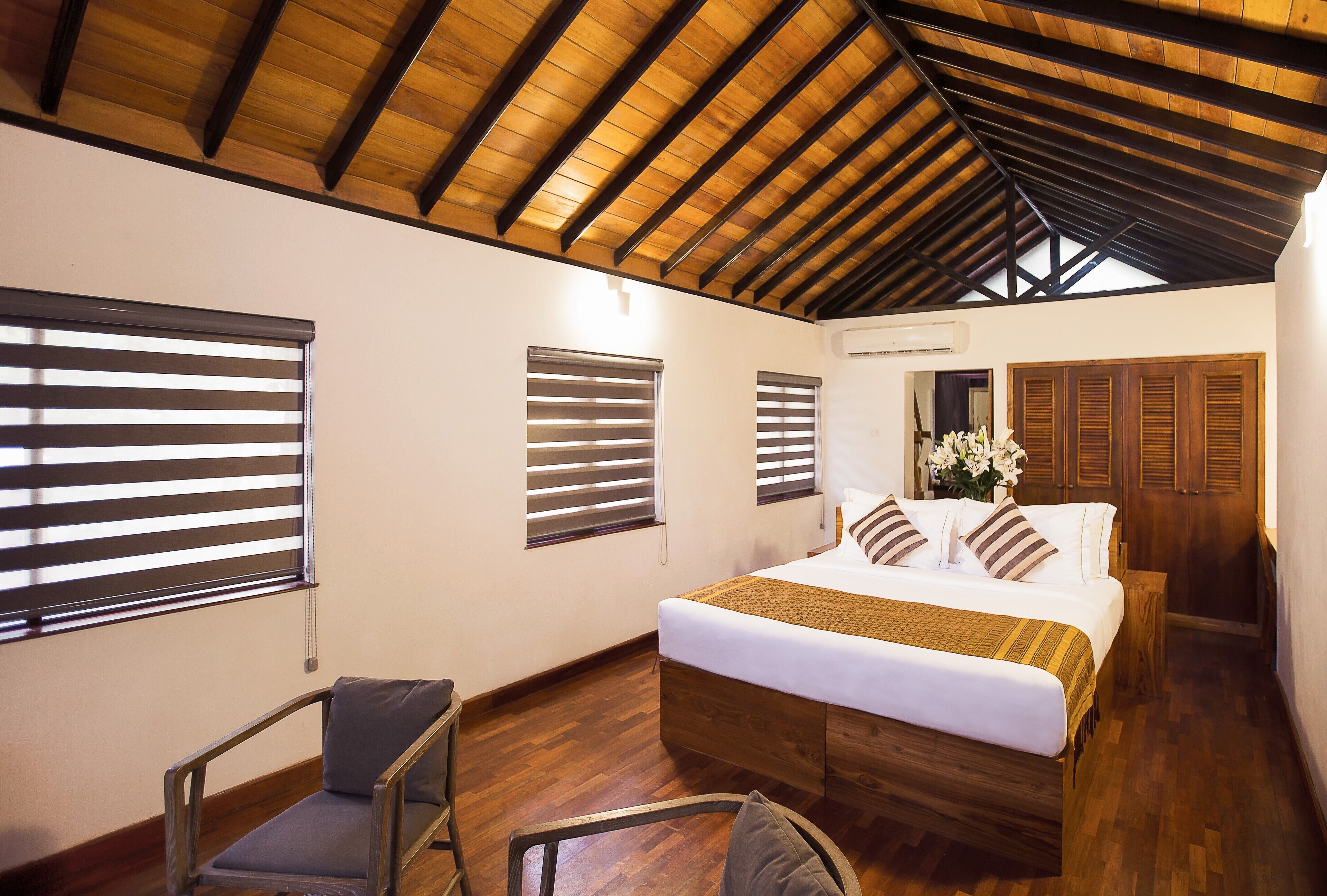 A bedroom with a wooden ceiling, a bed with white bedding, wooden shutters, and a chair.