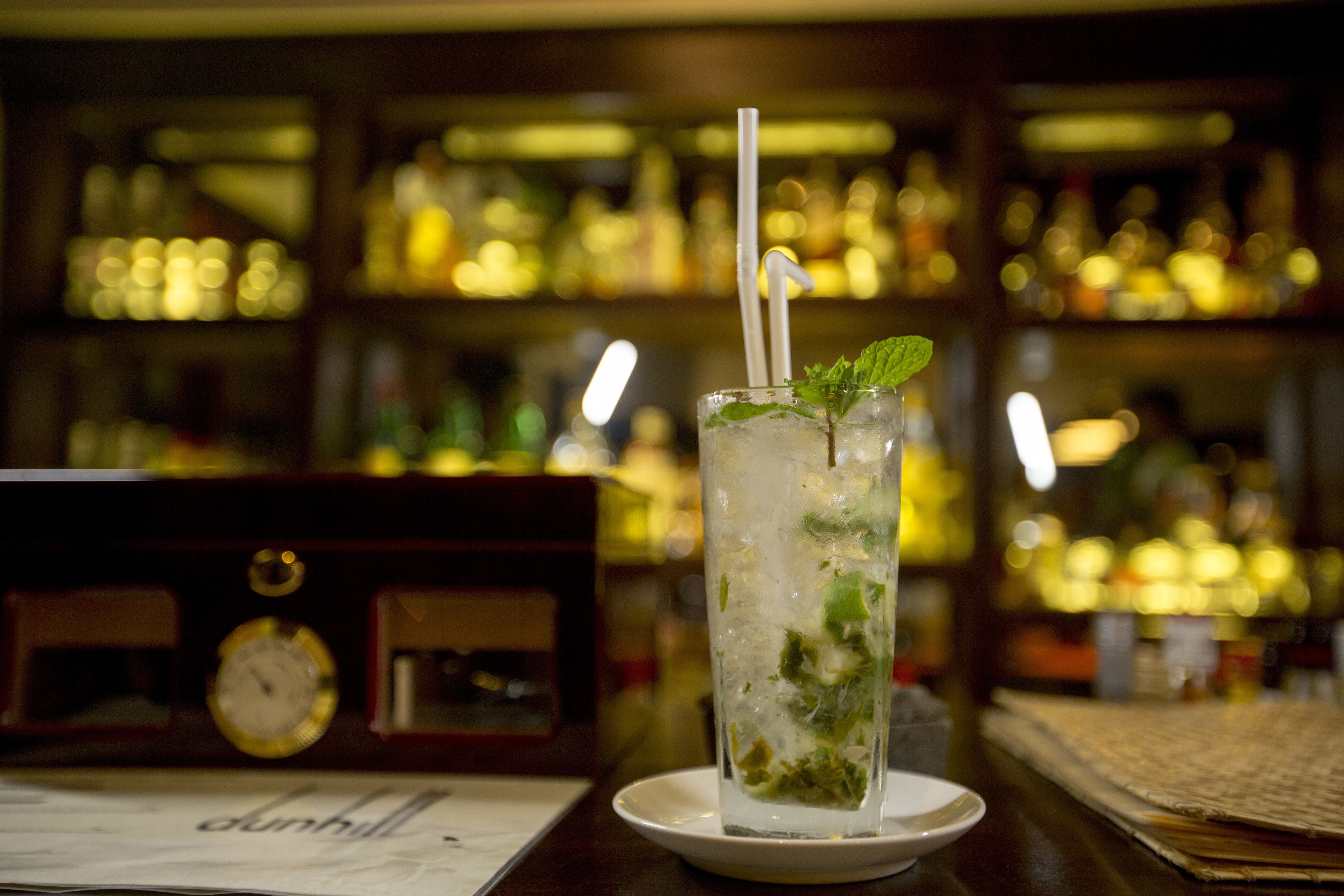 Ambiente de bar com um copo de bebida gelada decorado com hortelã, um relógio e um cardápio sobre a mesa.