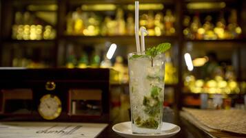 Ambiente de bar com um copo de bebida gelada decorado com hortelã, um relógio e um cardápio sobre a mesa.